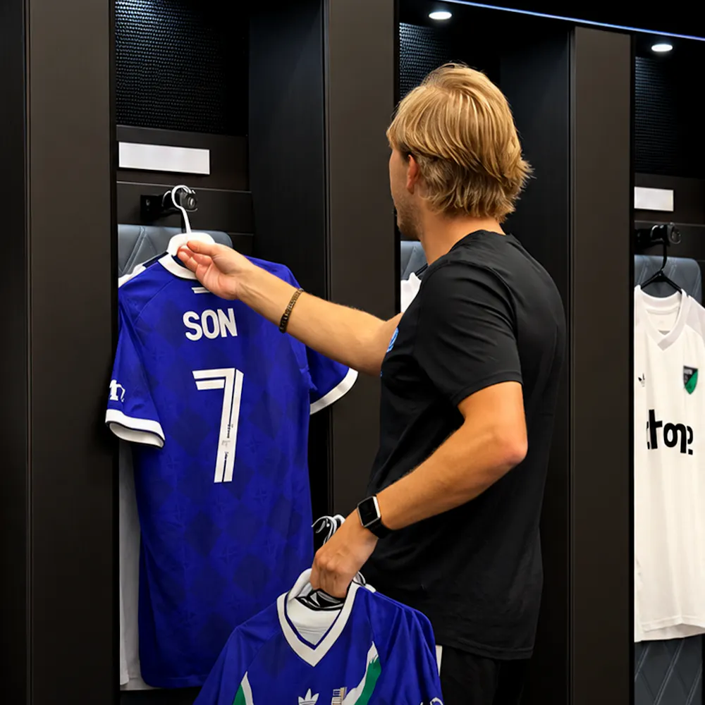 A man arranges a blue soccer jersey with "Son" and the number 7 in a locker room, holding another jersey in his left hand.
