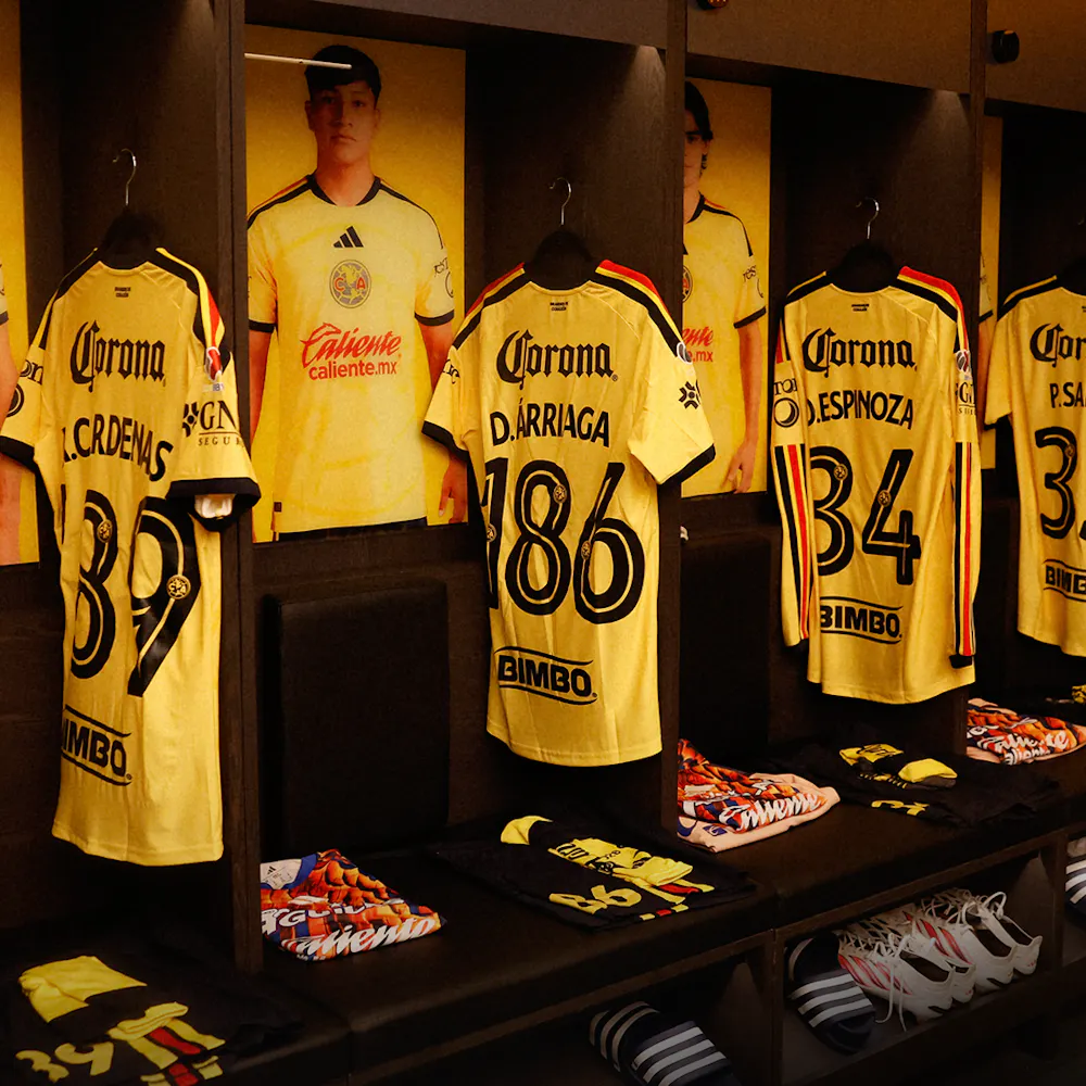 Yellow soccer jerseys with player names and numbers hang in a locker room. Beneath them are neatly folded clothes and sports shoes.