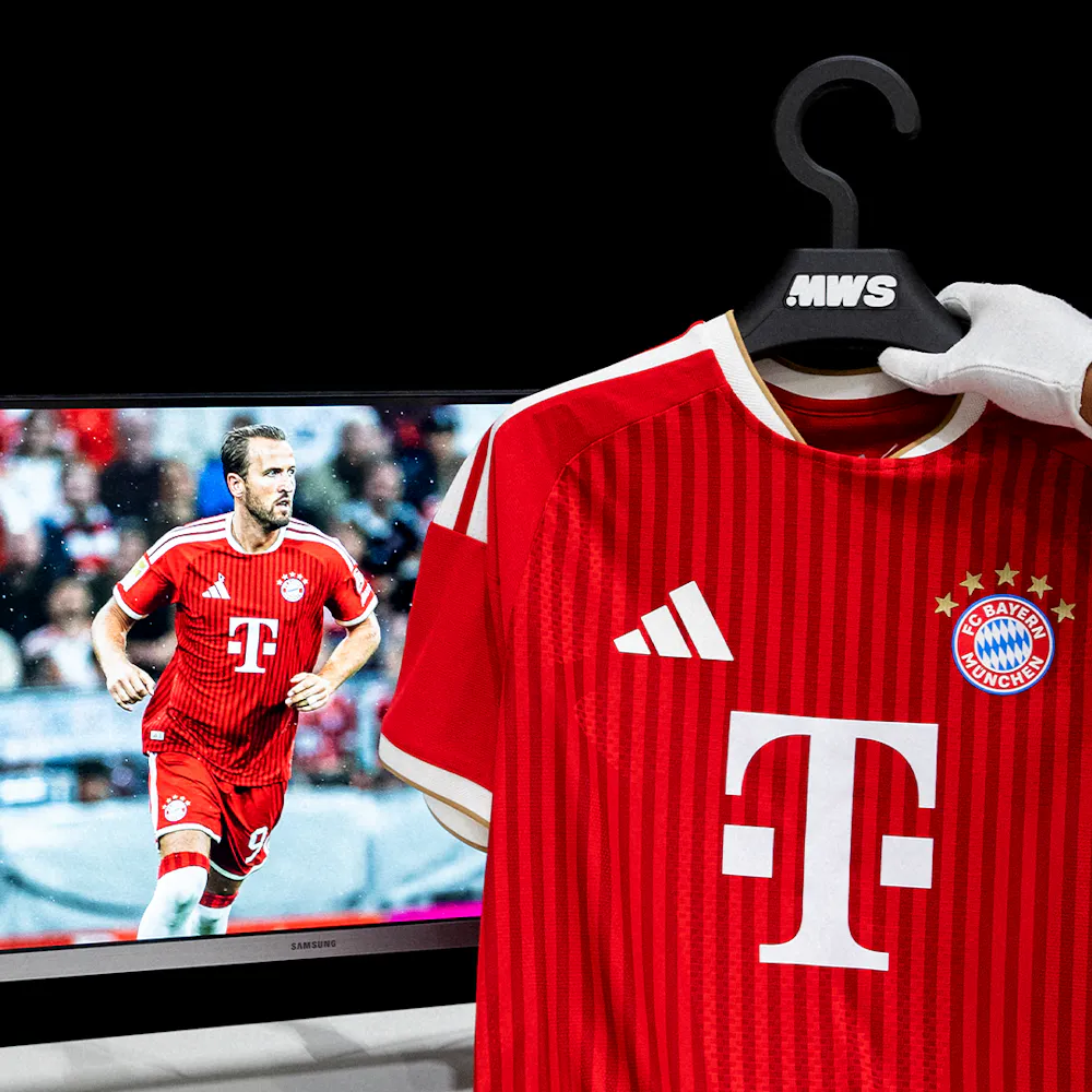 A Bayern Munich jersey on a hanger is held in front of a screen showing a player wearing the jersey during a game.