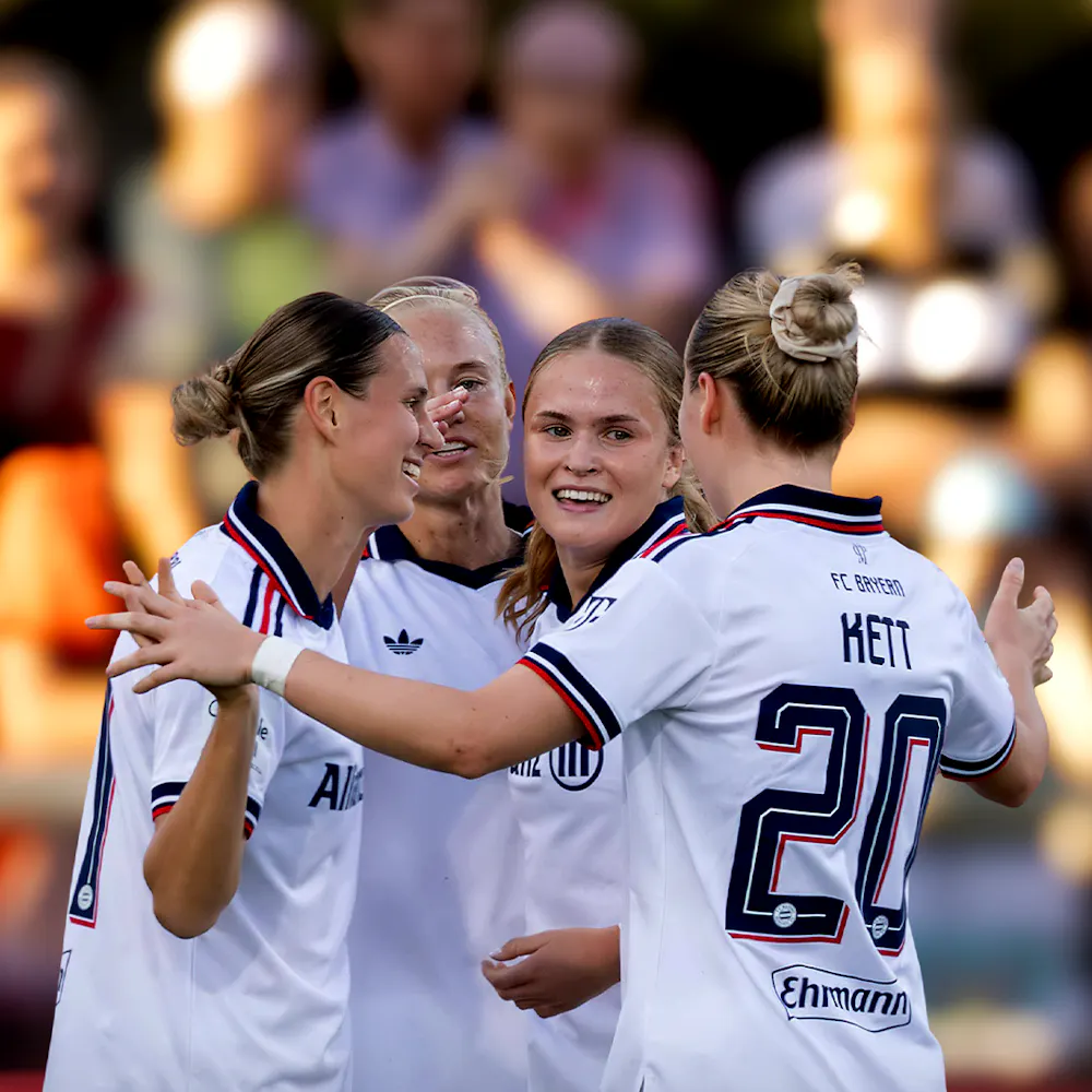 Four female soccer players in white uniforms, celebrating on the field, with blurred crowd in the background.