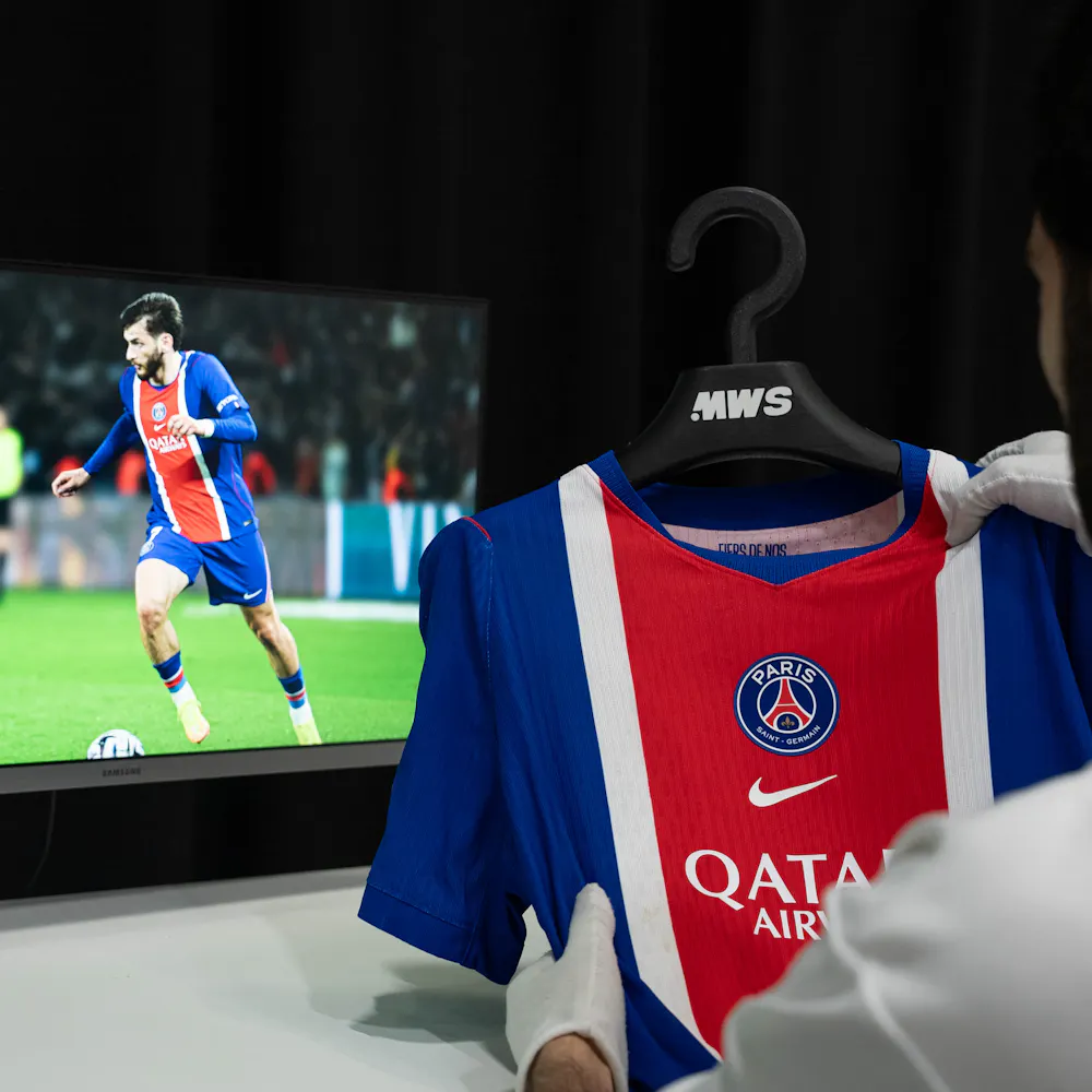 A person holds a Paris Saint-Germain football jersey near a screen showing a player in the same uniform on the field.