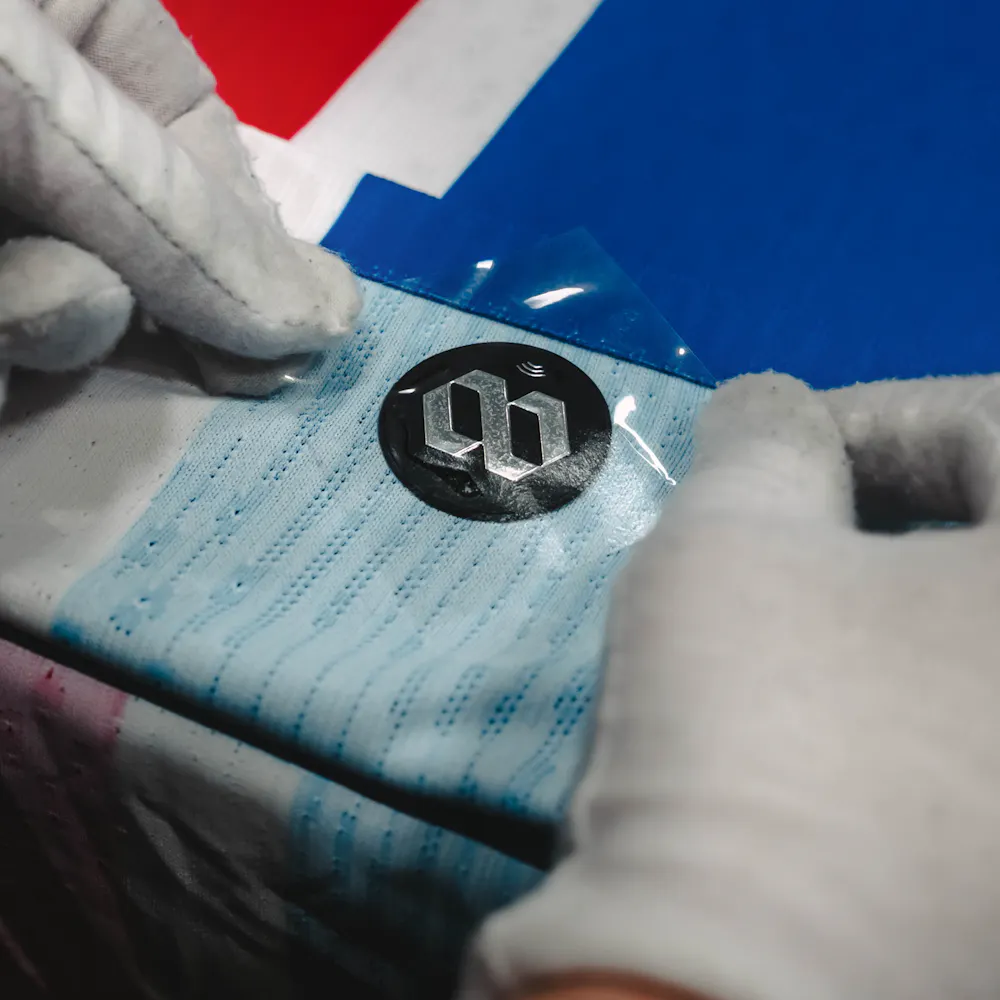 Gloved hands applying a blue protective film over a black circular logo on a textured fabric with red, white, and blue colors.