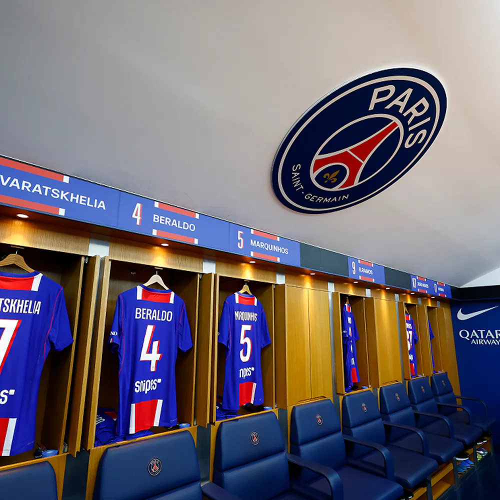 A football locker room featuring Paris Saint-Germain jerseys with names and numbers, blue and red design, and a Paris Saint-Germain emblem on the ceiling.
