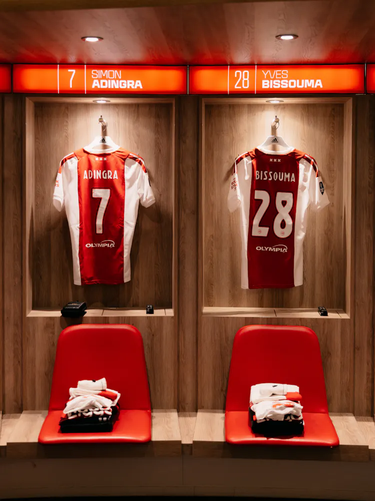 Ajax lockerroom shirts. Two red and white football jerseys hanging in a locker room, labeled with the names Adingra and Bissouma, with red chairs below.